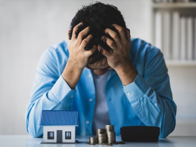 Man showing frustration beside house model and coins — representing mortgage stress and financial pressure.