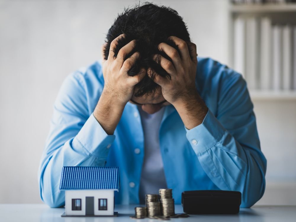 Man showing frustration beside house model and coins — representing mortgage stress and financial pressure.